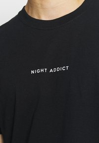 Juodas medvilninis marškinėlis su apvaliu kaklu. Krūtinėje yra balta spausdinta frazė "NIGHT ADDICT". Lygus audinio paviršius.