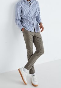 Páskovaná modro-bílá košile na knoflíky, olivově zelené kalhoty a bílé tenisky s gumičnou podr ážkou. Model pózuje s rukama v kapsách.