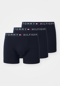 Três pares de boxers pretos Tommy Hilfiger com faixas elásticas assinadas, empilhados em disposição escalonada sobre um fundo branco.