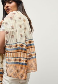 Lightweight, patterned scarf in cream with floral motifs and striped detailing in brown, blue, and gold. Features intricate edging and soft texture.