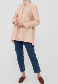 Camicia color pesco chiaro con colletto e maniche lunghe, vestibilità rilassata e orlo curvo, abbinata a jeans blu a gamba dritta e mocassini marroni.