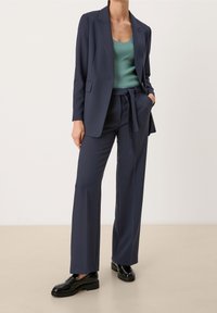 Tmavě modré sako s hladkou texturou, doplněné o sladěné kalhoty na míru a žebrovaný zelený top. Černé lakované mokasíny dokončují vzhled.