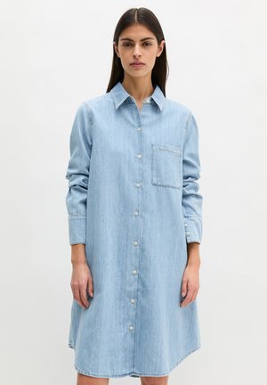 Frau mit glatten dunklen Haaren trägt ein hellblaues Denim-Kleid mit langen Ärmeln, Knopfleiste vorne und einer Brusttasche, steht vor weißem Hintergrund.