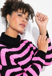 Mustat ja vaaleanpunaiset raidoitetut pooloneuleet, joissa on pehmeä ja teksturoidun näköinen neulos. Mallilla on yllään suuret hopeiset renkaat, ja hän poseeraa lähikuvassa.