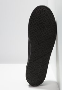 Semelle de chaussure noire avec motif géométrique texturé, appuyée contre un mur blanc sur une surface blanche.
