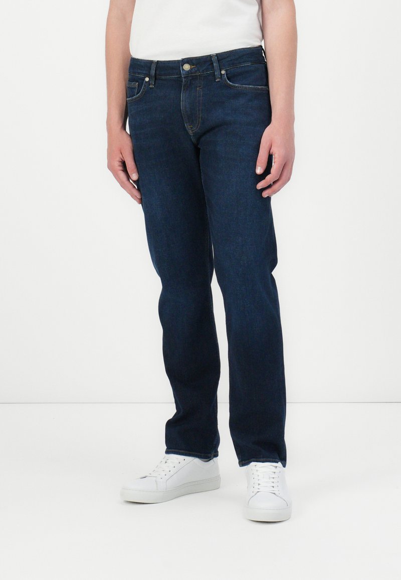 Dark blue denim jeans with a straight leg cut, featuring five pockets, a button closure, and contrasting yellow stitching. Paired with white sneakers.