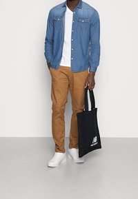 Jeansskjorta med knappar, vit T-shirt, beige chinos, vita sneakers, svart toteväska med vitt "New Balance"-logga; avslappnad och funktionell outfit.