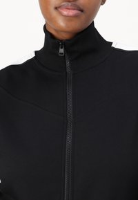 Schwarze Jacke mit Reißverschluss und hohem Kragen, gerippte Struktur und weiße Akzentstreifen auf den Schultern. Verfügt über einen Frontreißverschluss und glatten Stoff.