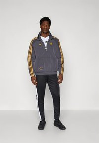 Grå jacka med dragkedja, gula ränder, broderad logotyp och framficka. Matchas med svarta byxor med vita sidoremmar och svarta skor.