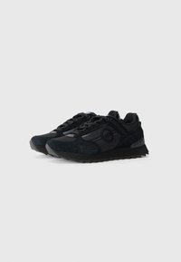 Baskets de sport noires en daim et en matériaux synthétiques, avec une semelle texturée, des lacets et un détail en forme de logo rond sur le côté.