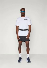 Hombre de pie de frente, vistiendo una camiseta de running blanca de Adidas, pantalones cortos negros, gafas de sol deportivas azules, calcetines blancos y zapatillas de running negras.