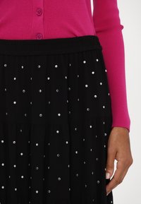 Rosa ribbad cardigan med runda knappar, kombinerad med en svart volangkjol prydd med silverfärgade cirkulära dekorationer.