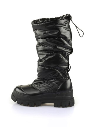 Personne portant des bottes matelassées noires, une mini-jupe noire et une veste noire, debout sur un trottoir urbain ensoleillé près d'un bâtiment blanc.