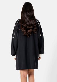 Vrouw met lang golvend donker haar, gekleed in een losse zwarte jurk met lange mouwen met witte manchetten en witte bies op de mouwen, staand met de rug naar de kijker gekeerd.