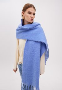 Lyseblå uldsjaal med en blød tekstur, der har frynser i kanten og en draperet design, båret over en cremefarvet top og afslappede jeans.