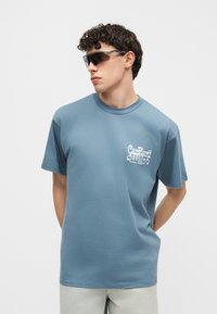 Junger Mann trägt blaues Grafik-T-Shirt und dunkle Sonnenbrille, steht mit den Händen hinter dem Rücken und schaut nach links vor einem einfarbigen Hintergrund.