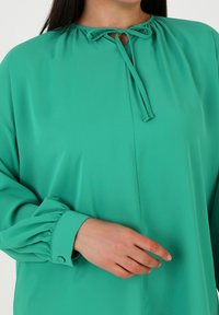 Türkise Bluse aus glattem Stoff mit gerafftem Ausschnitt und Bindedetail. Hat lange Ärmel mit gerafften Manschetten und Knopfakzenten.
