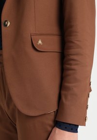 Brun kavaj med en gyllene monogramdetalj på fickan, med en slät textur och en skräddarsydd passform. Gyllene knappar vid ärmslutet ger en accent.