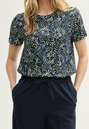 Kobieta w granatowej bluzce z krótkim rękawem ozdobionej zielonymi, niebieskimi i białymi abstrakcyjnymi plamami oraz granatowych spodniach z troczkiem, z rękami w kieszeniach.