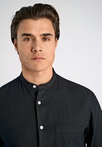 Camicia nera con colletto alla coreana, dotata di taschino frontale e bottoni bianchi su un tessuto texturizzato. Il modello posa direttamente di fronte alla telecamera.