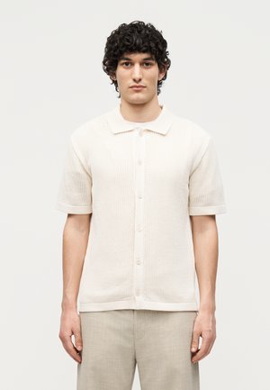 Mand med krøllet hår iført en kortærmet, knaplukket, off-white striktrøje og lyse beige bukser, stående mod en ensfarvet baggrund.