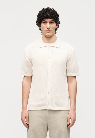 Uomo con capelli ricci che indossa una maglietta a maniche corte, abbottonata, color panna e pantaloni beige chiaro, in piedi davanti a uno sfondo semplice.
