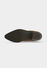 Chaussure en cuir marron à bout pointu vue de la semelle noire, montrant un petit talon avec une semelle texturée sur un fond blanc uni.