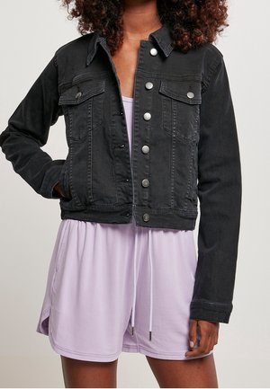 Woman wearing a black denim jacket over a light purple dress with drawstrings, standing with one hand in jacket pocket.