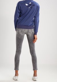 Modrá mikina s velkým lesklým logem na zadní straně, spojená se šedými úzkými džínami. Bílá teniska dotváří outfit, pohled zezadu.