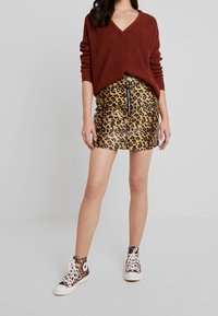 Nainen, jolla on päällään ruskea V-poolopaita, leopardikuvioinen minihame, jossa on vetoketju edessä, ja samasta leopardikuvioisesta kankaasta valmistetut korkeavartiset tennarit, seisoo valkoista taustaa vasten.