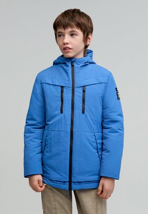 Chaqueta aislante azul con capucha, que cuenta con dos bolsillos en el pecho con cremallera, detalles de cremallera en negro y un acabado de tela texturizada.