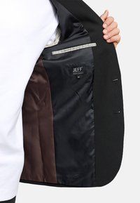 Schwarze Blazer-Innenausstattung mit braunem und schwarzem Satinfutter, versehen mit einem Etikett, das "JEFF, Urban Tailoring, Größe 50" liest, und einem gemusterten Akzent.