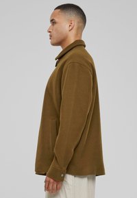 Brown fleece jacket with a collar, buttoned cuff details, and side pockets, displayed from the side against a neutral background.