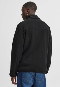 Giacca in pile nera con colletto alto e maniche lunghe, caratterizzata da una texture morbida e dettagli della cucitura sul retro. Abbinata a jeans blu.