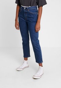 Vaqueros azules de cintura alta con pierna recta y dobladillo cónico, combinados con zapatillas altas blancas sobre un fondo neutro.