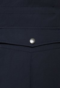 Granatowy materiał o gładkiej teksturze z kieszenią z przodu zapinaną na srebrny zatrzask. Widoczne szwy podkreślają kieszeń i krawędzie odzieży.