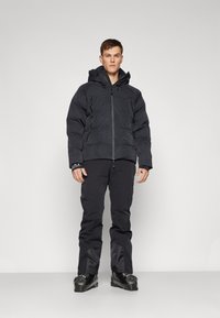 Jachetă de schi izolată, neagră, cu un model texturat în valuri, închidere cu fermoar, glugă și manșete ajustabile, asortată cu pantaloni de schi negri și bocanci negri.