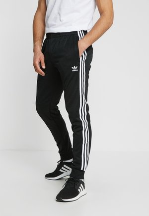 Man draagt zwarte Adidas trainingsbroek met witte strepen langs de zijkanten en bijpassende zwarte Adidas sneakers met witte strepen.