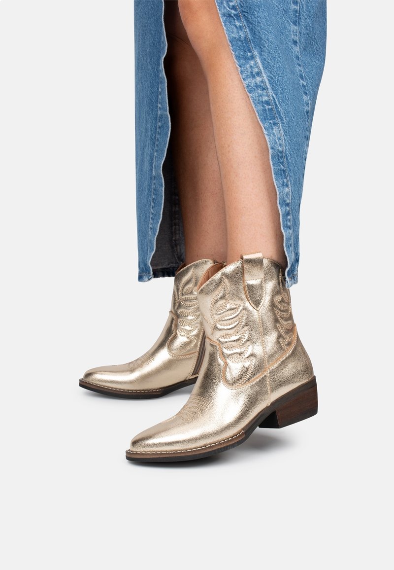 Zlaté kovové kovbojské boty s špičatou špičkou, ozdobným prošíváním a nízkým dřevěným podpatkem, nošené s džínovou sukní s bočním rozparkem.