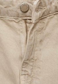 Tejido de mezclilla beige con cierre de botón de latón y cremallera metálica visible. Presenta una textura fina y sutilezas en los detalles de costura.