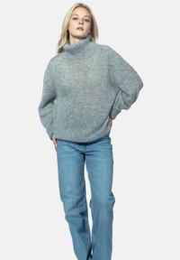 Jasnoszary, oversize sweter z wysokim kołnierzem i prążkowanymi mankietami, wykonany z miękkiego, teksturowanego materiału, w zestawie z jasnoniebieskimi dżinsami o prostych nogawkach.