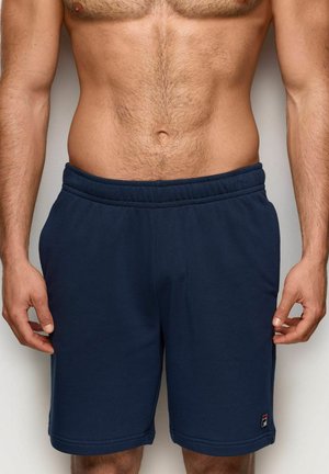 Mężczyzna z gołym torsem, ubrany w granatowe spodenki sportowe z elastycznym pasem i małym logo na dolnej lewej nodze, stojący z rozluźnionymi rękami.