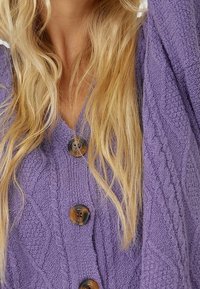 Paarse gebreide cardigan met een gestructureerd ontwerp, voorzien van grote bruine knopen en een V-hals. Lange mouwen reiken voorbij de pols.