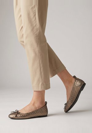 Person trägt beige Hose und metallisch gesteppte Ballerinaschuhe und läuft auf einem schlichten hellen Hintergrund.