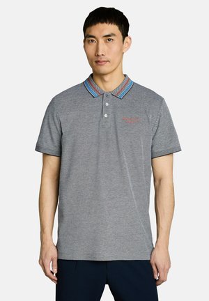 Homme portant un polo gris avec un col rayé bleu et rouge et un pantalon foncé, debout devant un fond clair uni.