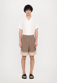 Camicetta bianca a maniche corte, pantaloni corti marroni a fantasia con orlo beige e sandali neri a ciabatta. Design semplice, tessuto leggero, vestibilità rilassata.