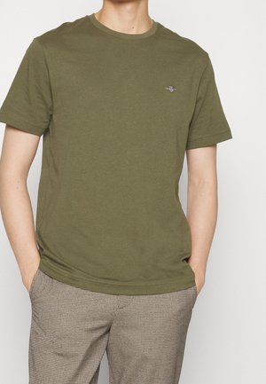 Osoba ubrana w oliwkowo-zielony t-shirt z krótkim rękawem i beżowe spodnie w kratę, ręce w kieszeniach, na jednolitym tle.