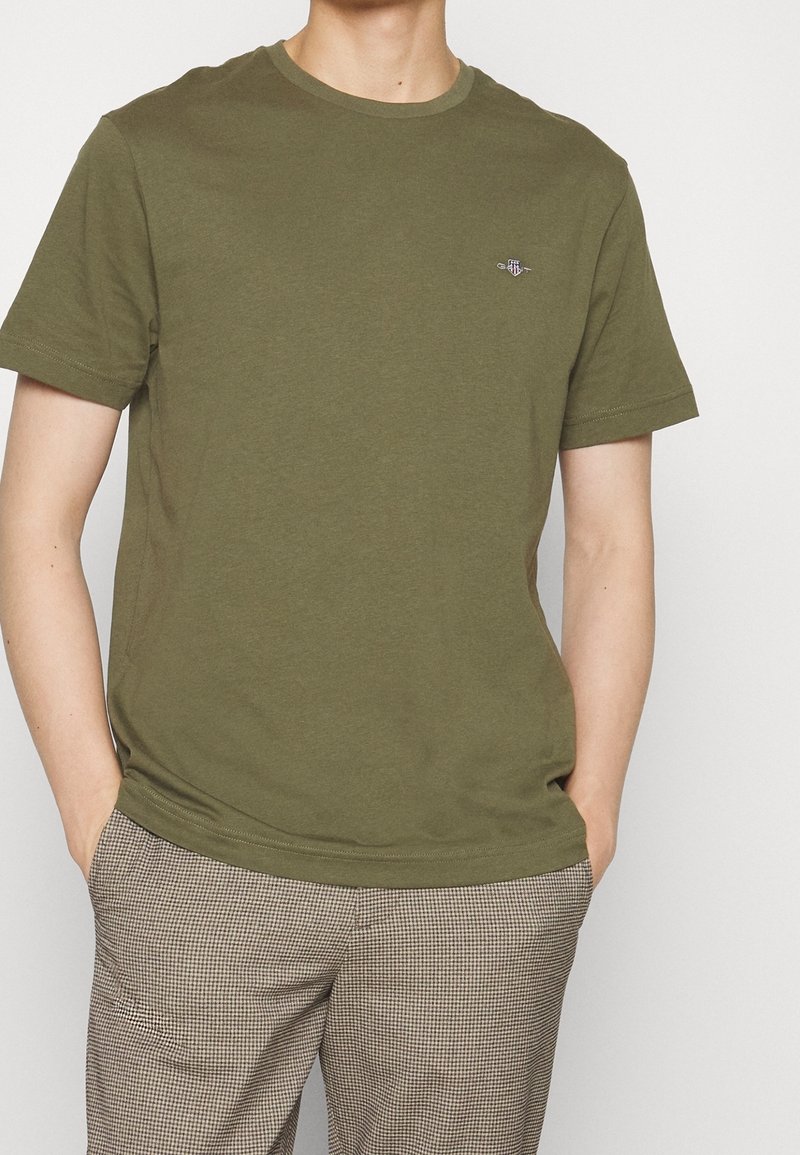 Personne portant un t-shirt manches courtes vert olive et un pantalon beige à carreaux, les mains dans les poches sur un fond uni.
