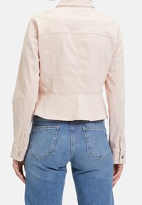 Světle růžová zkrácená bunda s jednoduchým designem, s knoflíkovými manžetami a viditelnými švy. Nepochybně nošena s modrými džíny, ukazujícími zadní a dolní detaily.