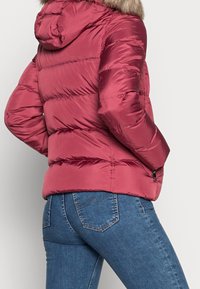 Czerwona, pikowana kurtka puchowa z kapturem wykończonym sztucznym futrem. Charakteryzuje się eleganckim krojem, kieszenią na zamek oraz błyszczącą, gładką fakturą materiału.